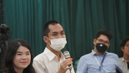 A photo of a participant, wearing white shirt and mask, and holding a mic. He seems to be asking some questions.