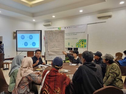 Beberapa peserta kegiatan tampak tenggelam dalam diskusi dengan satu sama lain. Mereka duduk di sebuah meja bundar. Sebuah papan berisikan poin-poin materi kegiatan berdiri di depan meja mereka.