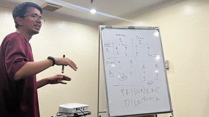 Rahman Imuda standing beside a board showing a prisoners' dilemma situation.